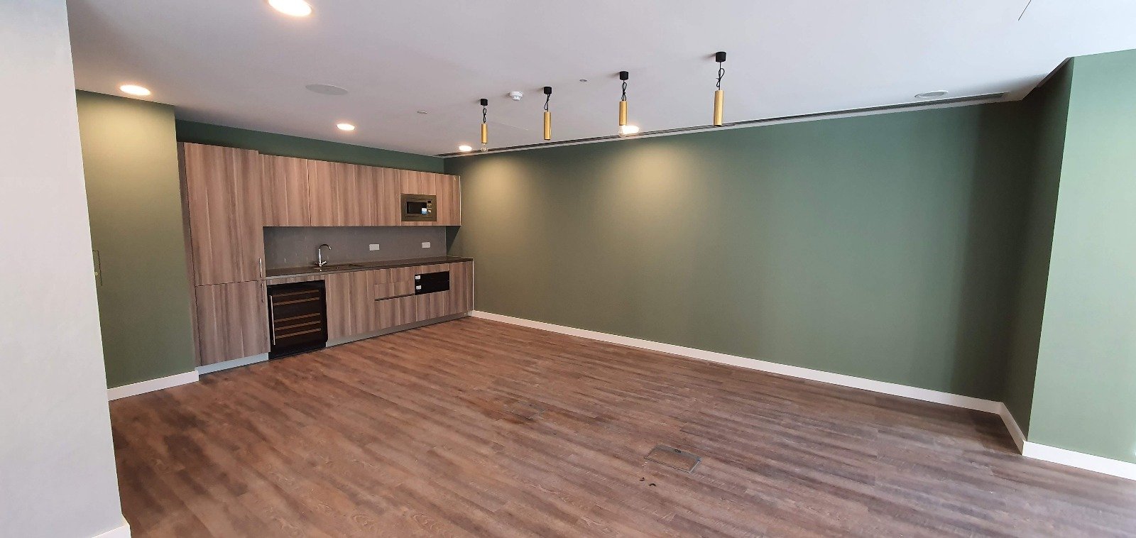 Empty room with wood flooring, green walls and a small kitchenette with wood cabinets.